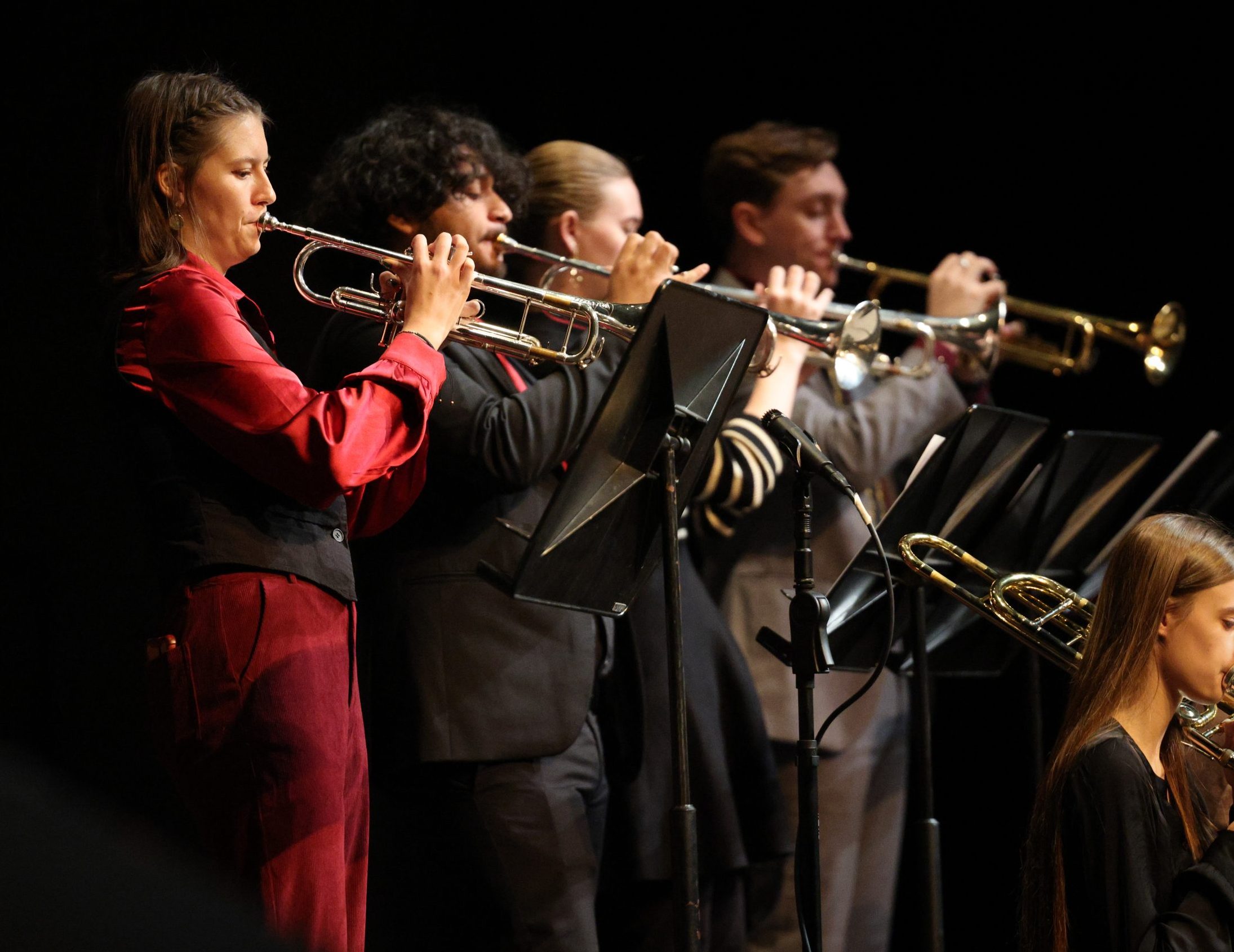 Four musicians stand in a row playing trumpets on stage, wearing formal attire. Sheet music stands highlight the focus on music, while a trombone player is seated in the lower right corner against a dark background.