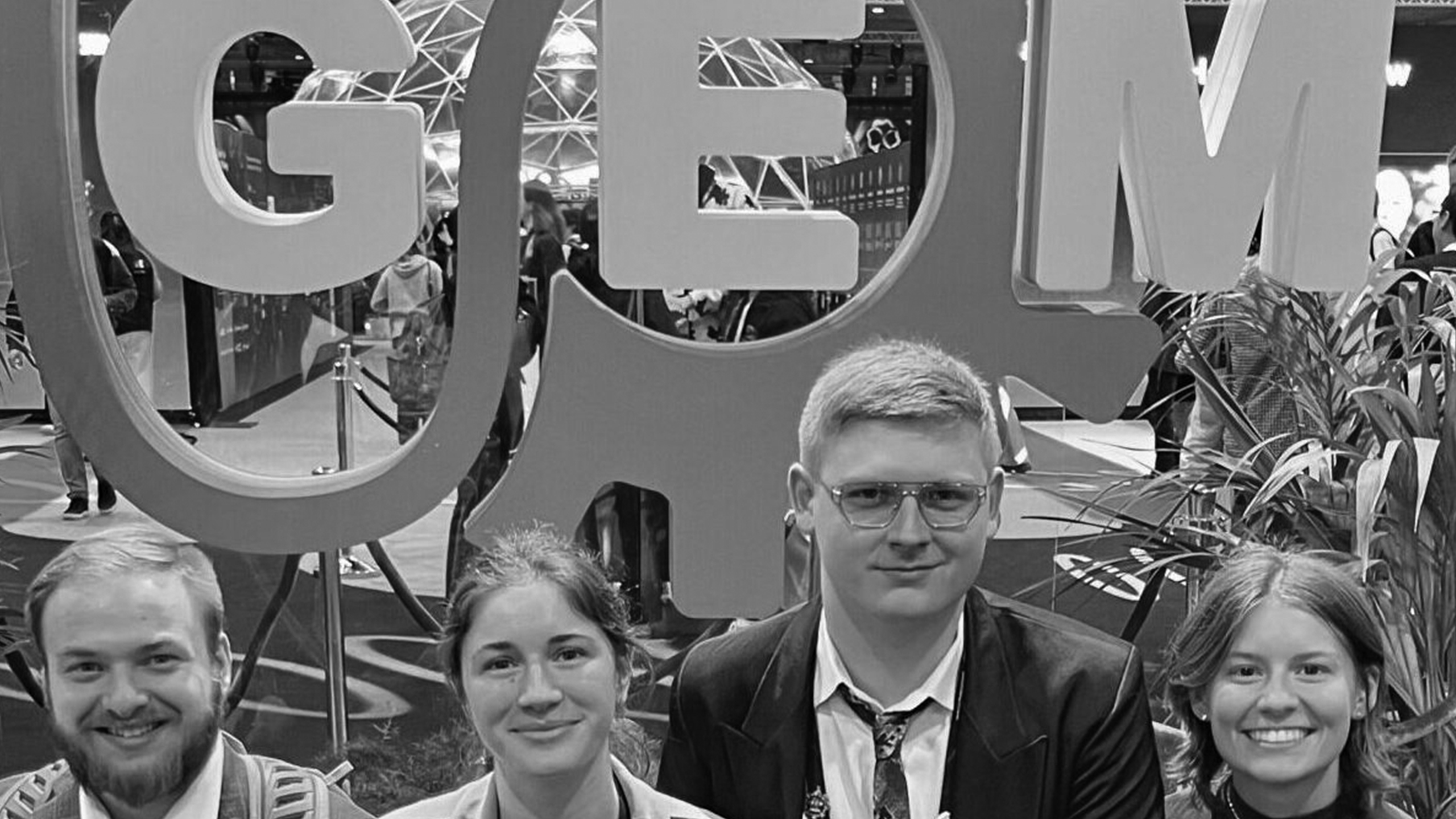 Four young adults, all pre-occupational-therapy students, pose and smile in front of a large GEM sign with a gear design at an indoor event. There are plants and other people in the background. The image is in black and white.