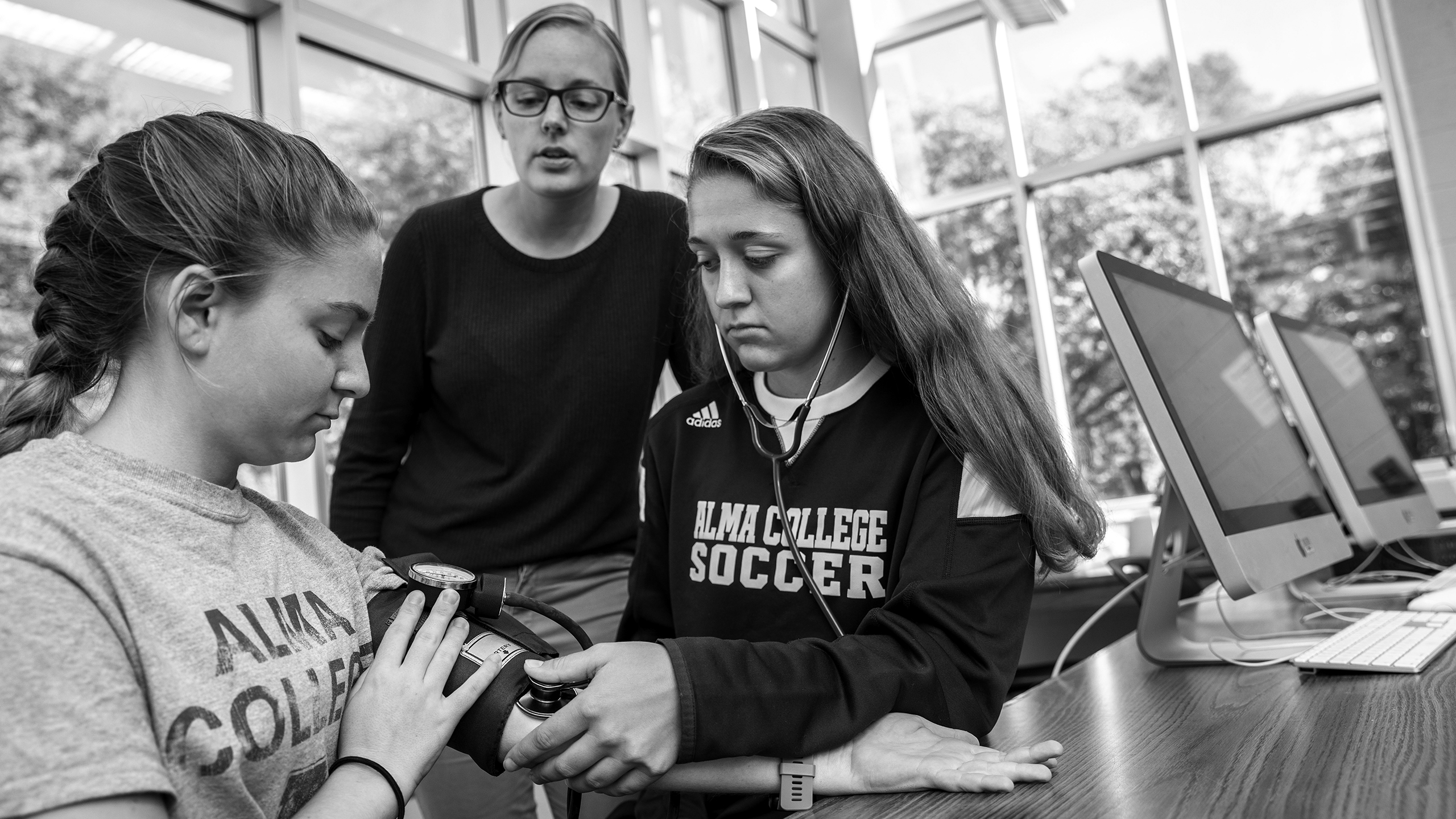 Two young women in a pre-occupational-therapy class practice taking blood pressure—one wearing an ALMA COLLEGE SOCCER shirt listens with a stethoscope, while a third person supervises in a bright classroom with computers.