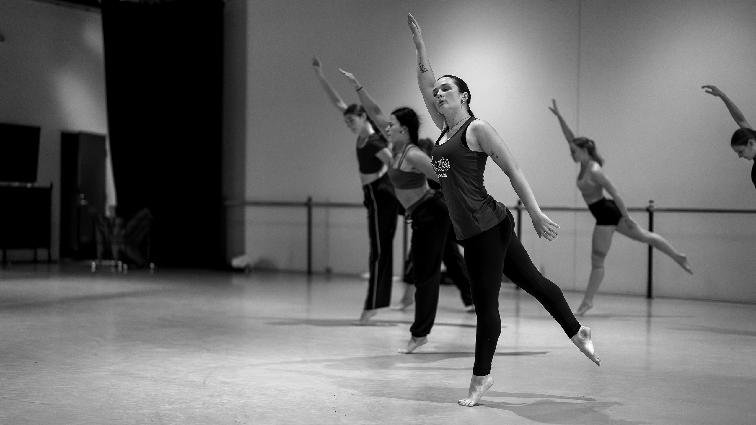 Four dancers practice in a studio, lifting one arm and one leg in a graceful pose. The black and white image highlights the motion, concentration, and pre-occupational-therapy benefits of their movements against a plain background.