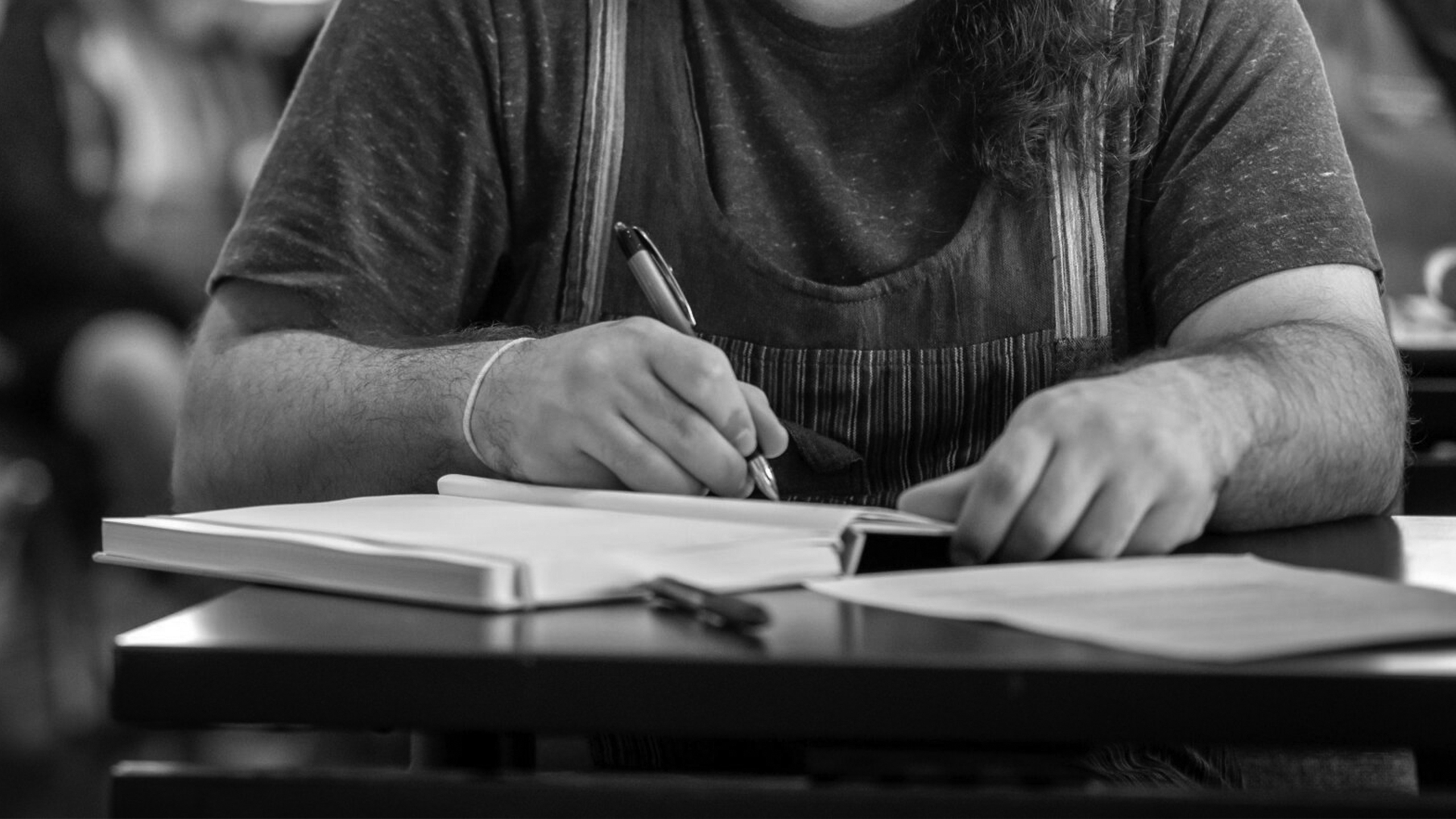A person sits at a desk, writing in a notebook with a pen, surrounded by papers. The black and white image focuses on the hands and torso, while other people appear blurred in the background.