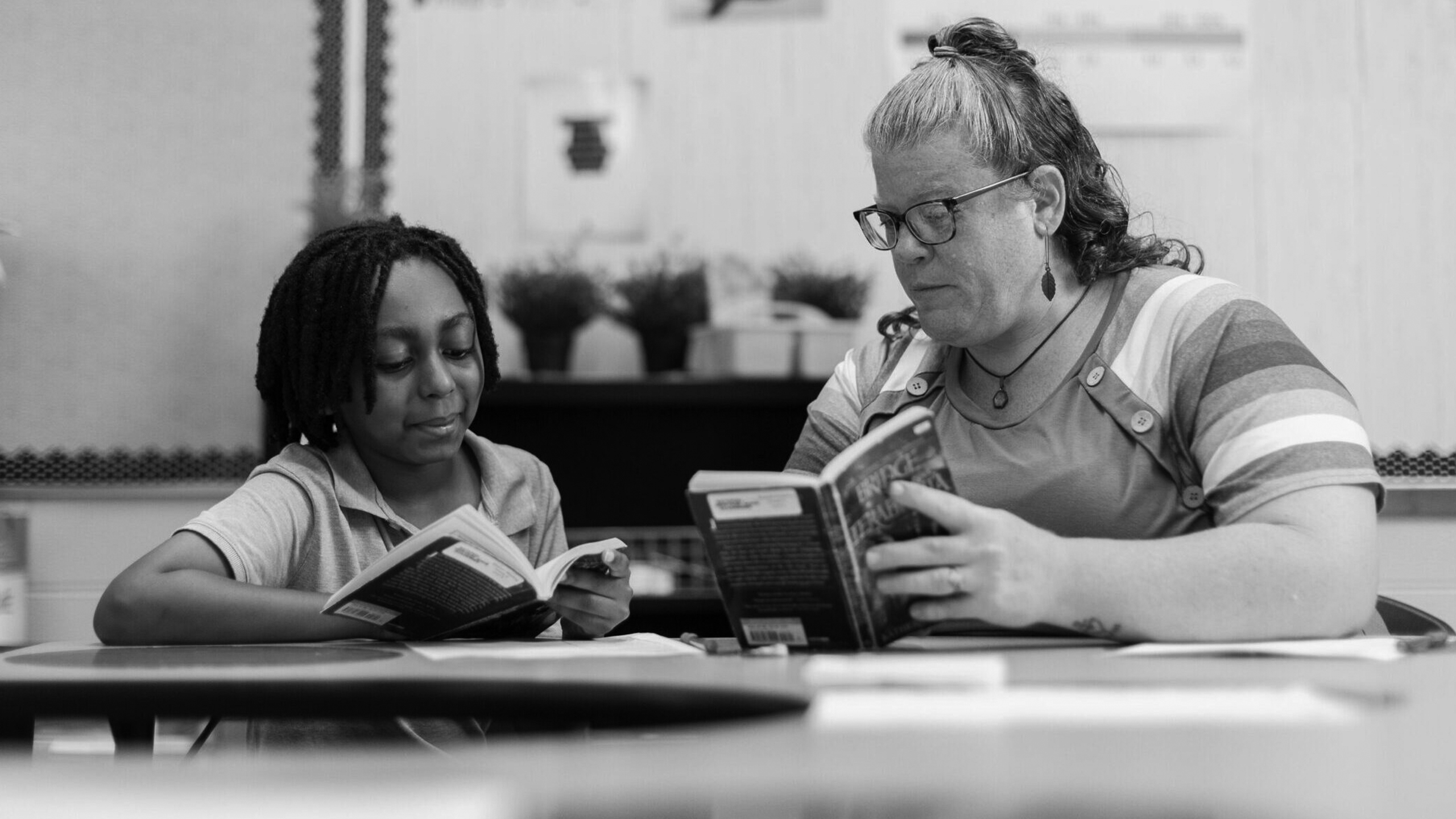 A pre-occupational-therapy student and a teacher sit together at a table, both reading books. The teacher, wearing glasses, looks at the focused student. Shelves and wall decorations can be seen in the classroom background.