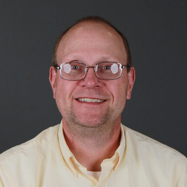 A middle-aged man with glasses, short hair, and a light yellow collared shirt smiles at the camera against a plain dark gray background.