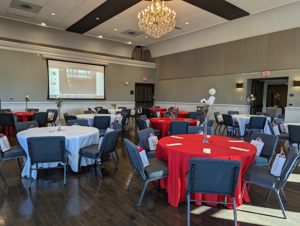 A banquet hall at Alma College's Opera House, with round tables covered in red and white tablecloths, gray chairs, elegant centerpieces, and a chandelier overhead. At the front, a projector screen. 