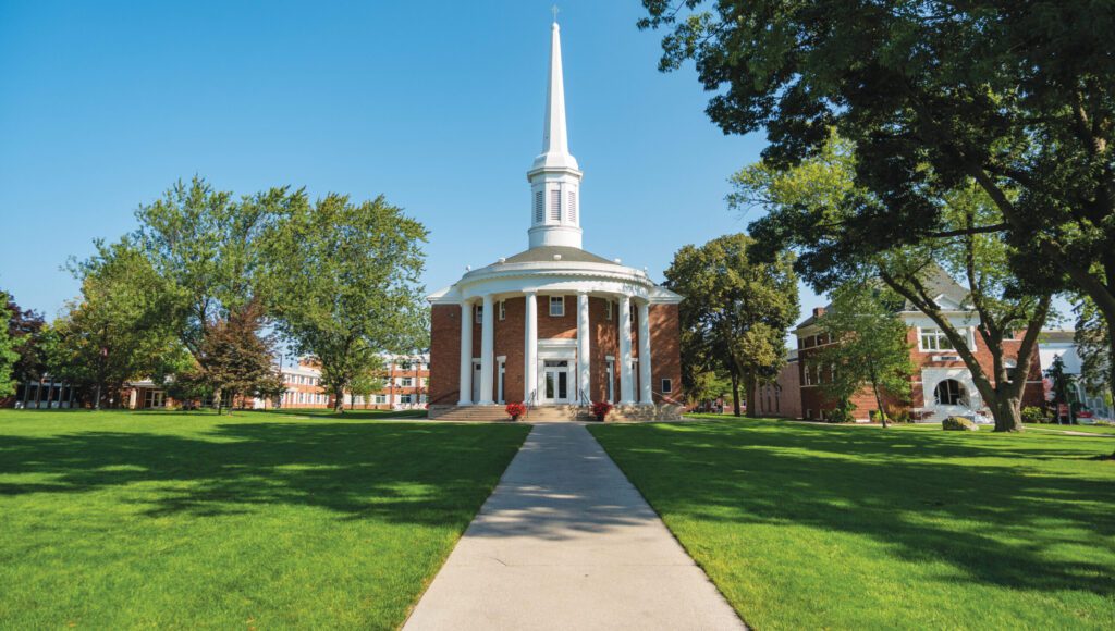 Alma College Andison Chapel in Summer stands at the end of a path, its brick exterior, tall white steeple, and columns surrounded by green lawns and trees under a clear blue sky.