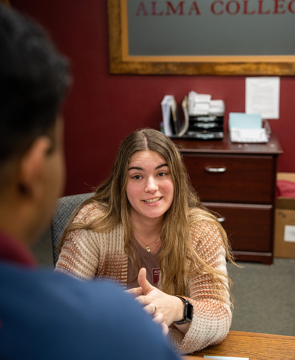 A student employee from admissions sitting behind a desk, explaining something to a student. The back of the student’s head is visible as they listen attentively to the explanation. The setting appears to be an office or admissions area.