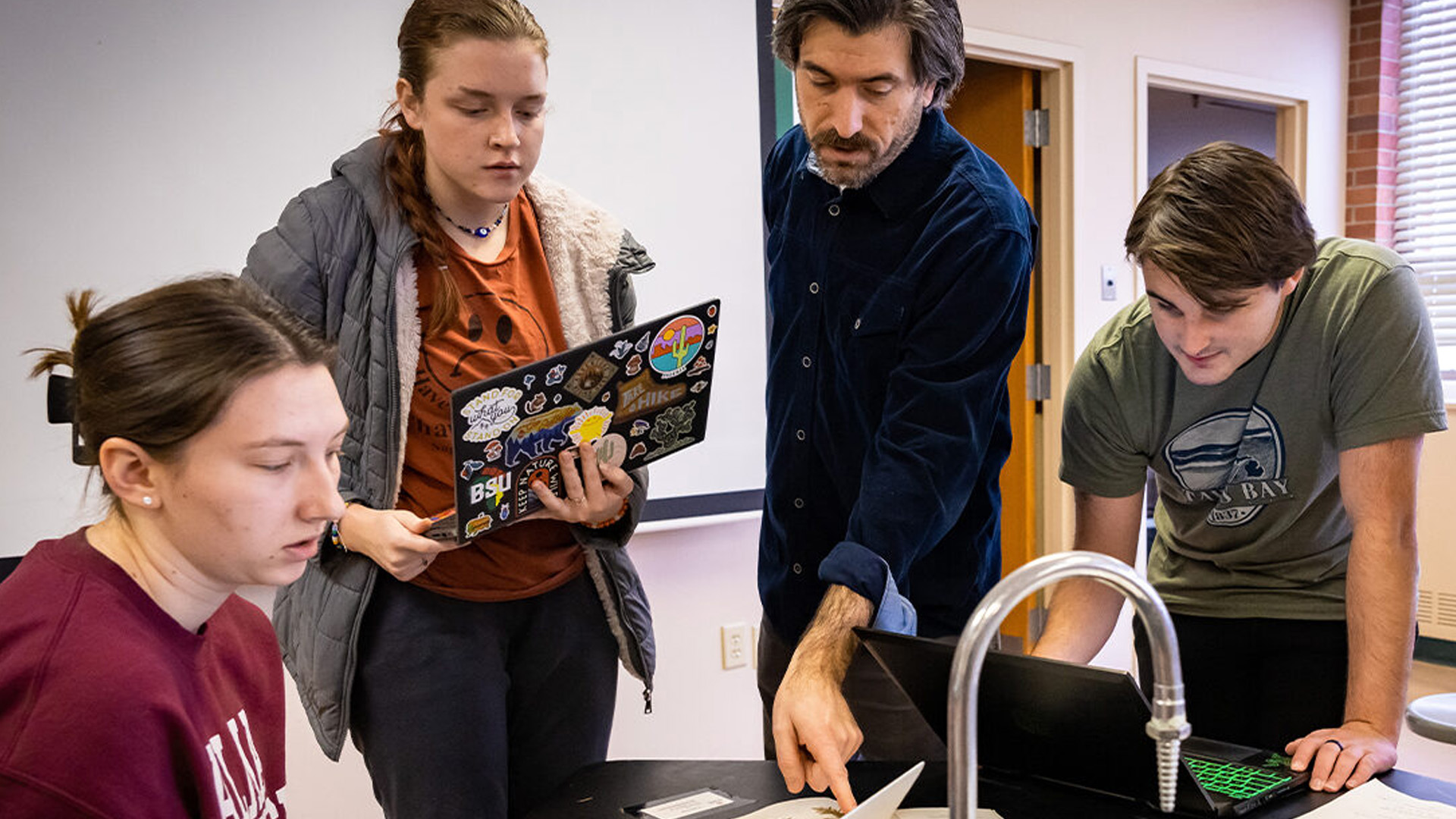 A professor pointing to a skeleton in the anatomy lab, with two students standing in front of him, learning. All three are wearing white coats as they engage in the lesson. The students are focused on the professor, who is explaining the anatomical features, with lab supplies visible in the background.