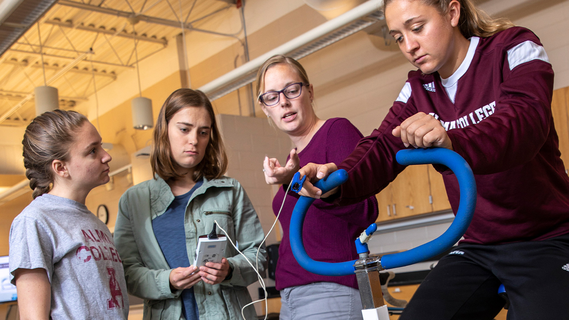 A professor pointing to a skeleton in the anatomy lab, with two students standing in front of him, learning. All three are wearing white coats as they engage in the lesson. The students are focused on the professor, who is explaining the anatomical features, with lab supplies visible in the background.