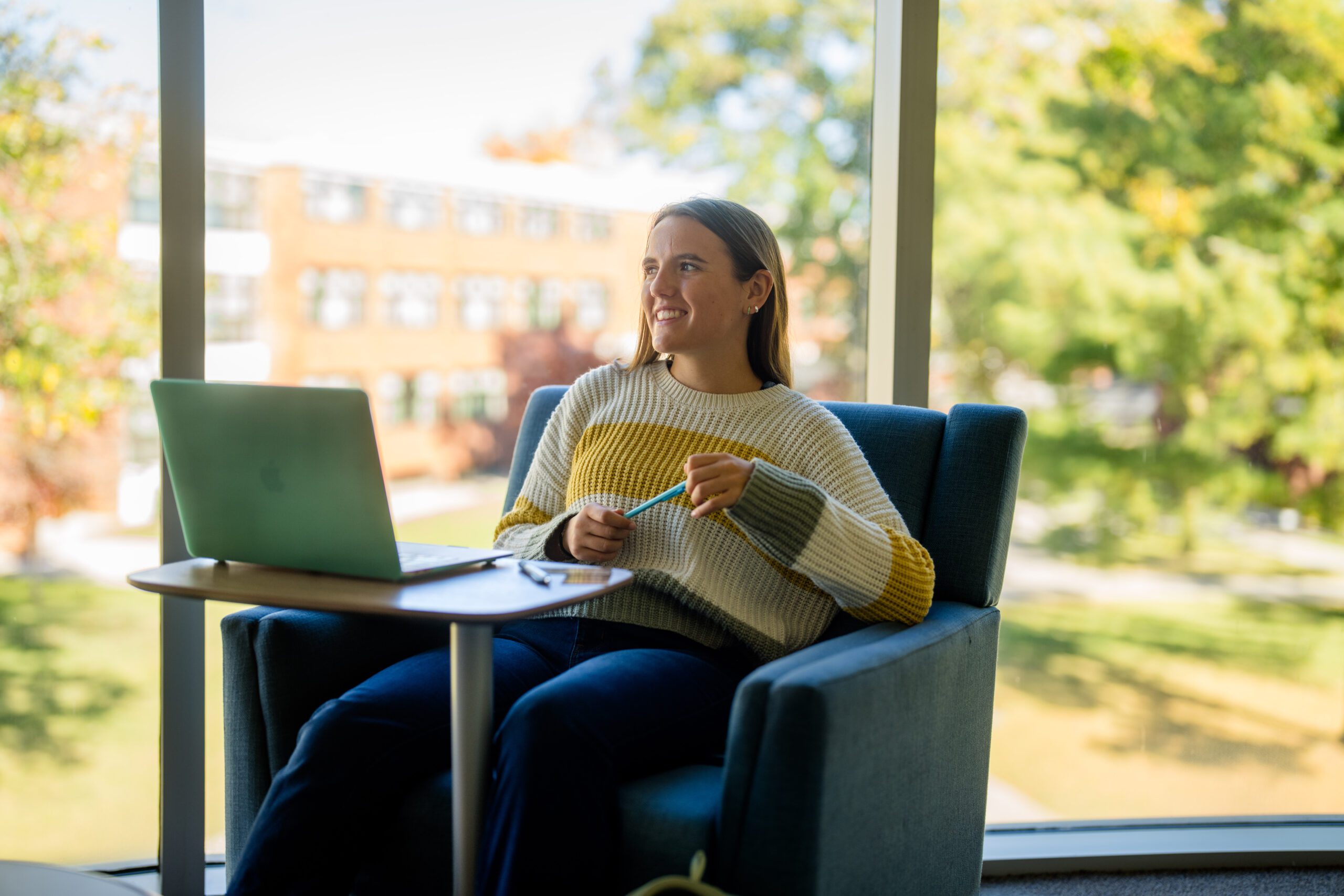 A young woman sits in a chair by a large window, smiling while holding a pencil and looking away from her open laptop on a small table. Bright greenery and buildings are visible outside.