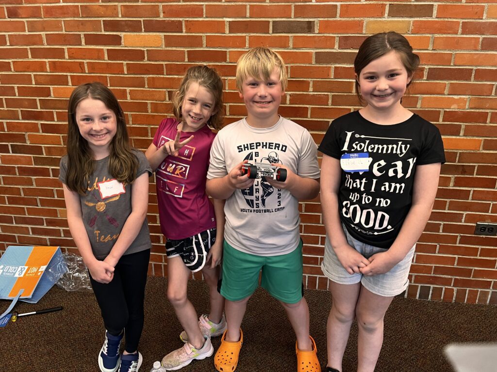 Four smiling children stand side by side in front of a brick wall at summer-at-alma-camps. The boy in the middle holds a small robot. They appear to be at a casual event, all dressed in comfortable, casual clothes.