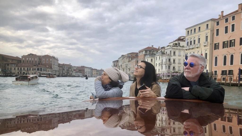 Three people are pictured on the water in Venice, Italy, looking out toward a scenic coastline.