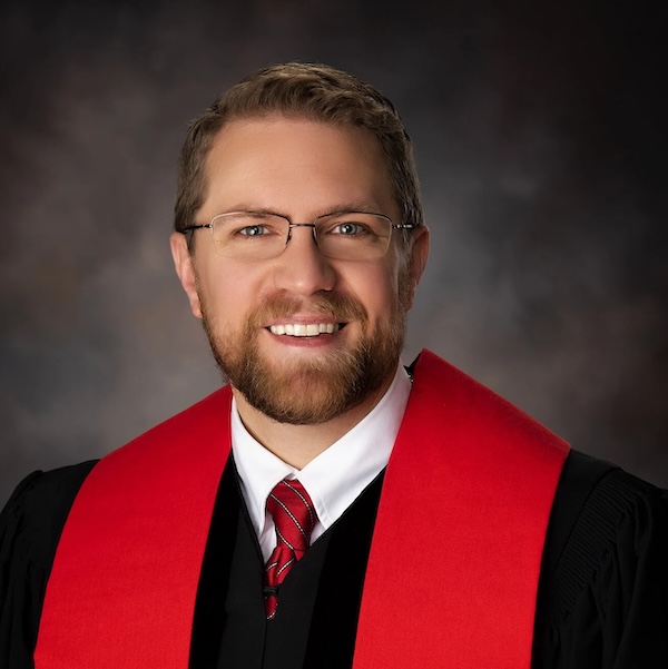 A man in religious clothing and glasses smiles for a posed photo.