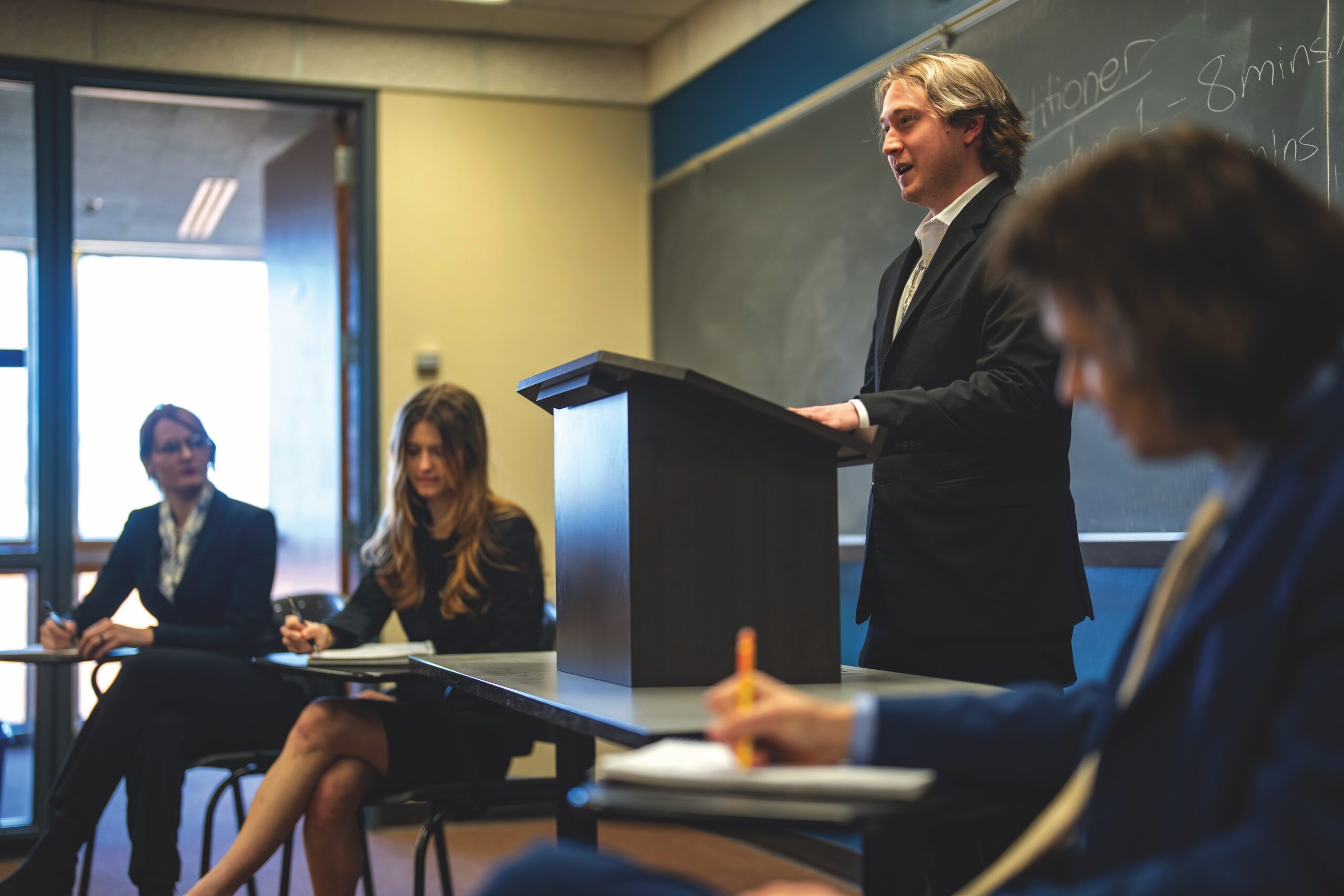 A student wearing a suit stands at a lectern in a college classroom, surrounded by similarly-dressed students.