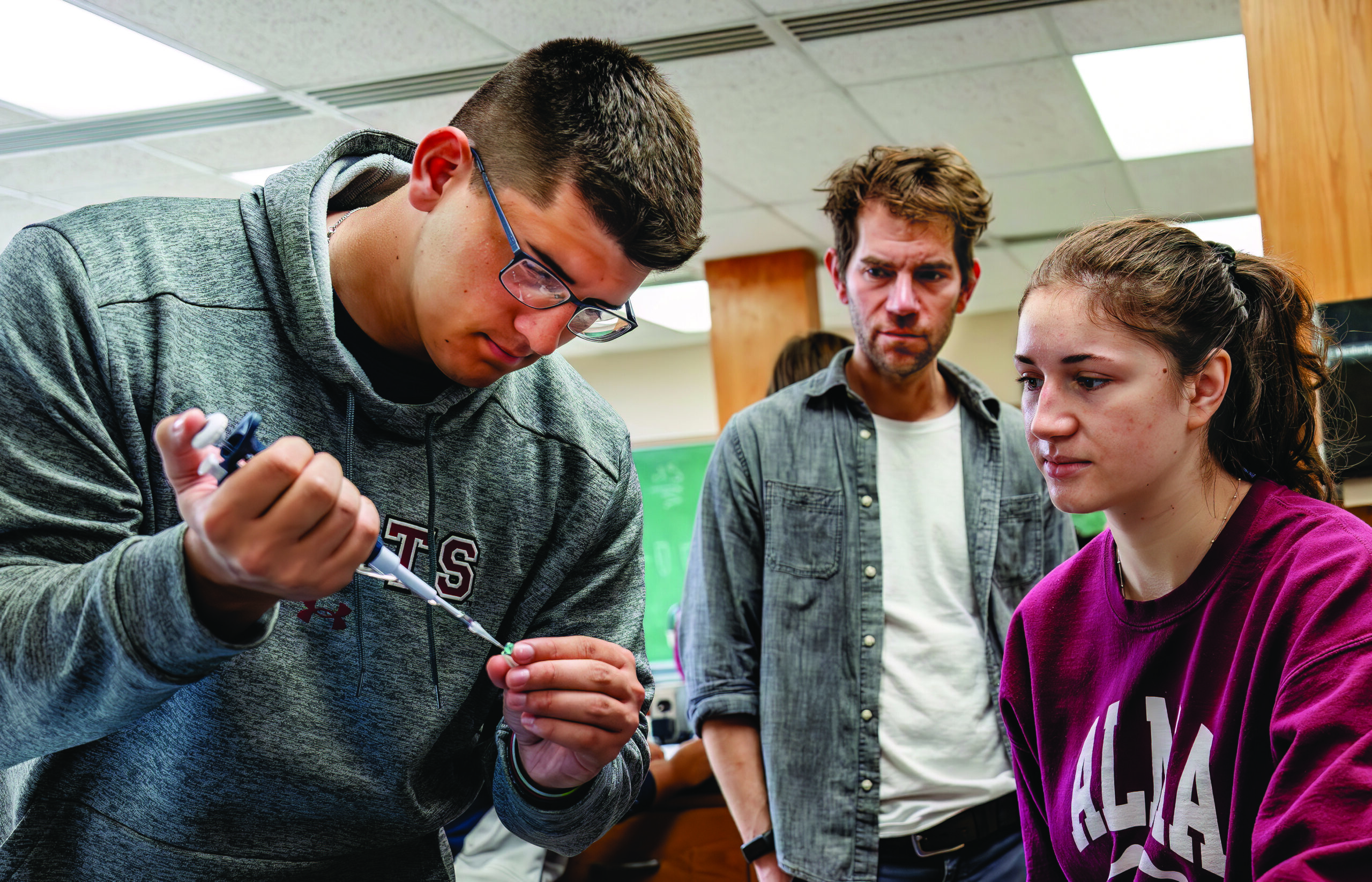 In a collegiate laboratory setting, a male student works with an eyedropper tool while a female student and male faculty advisor look on.