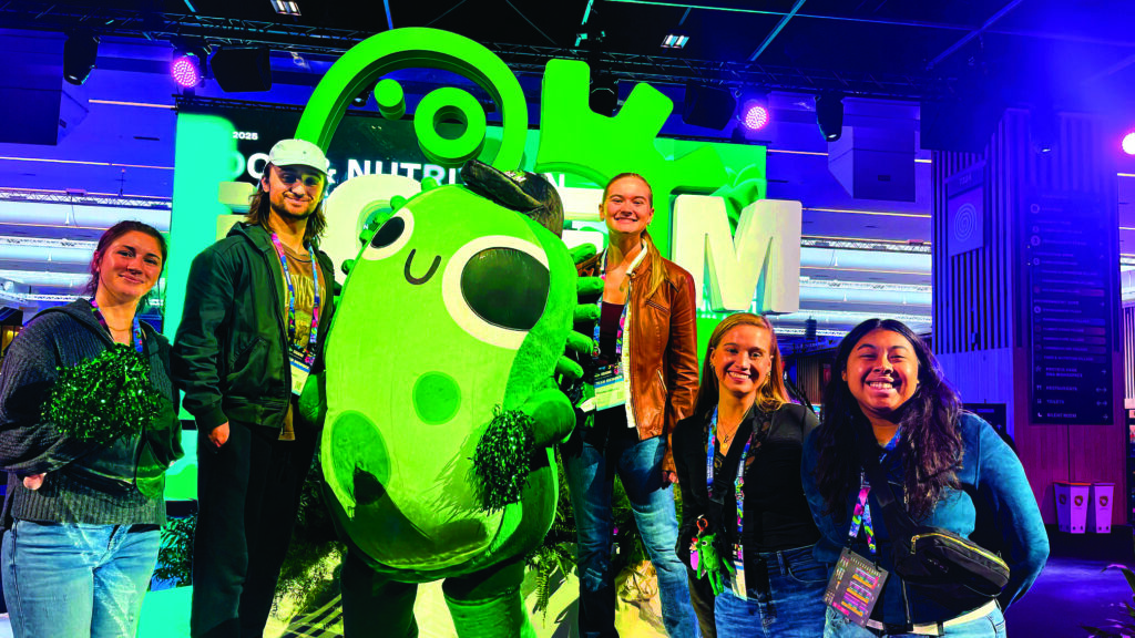 A group of students at a college conference surround themselves with a green mascot.