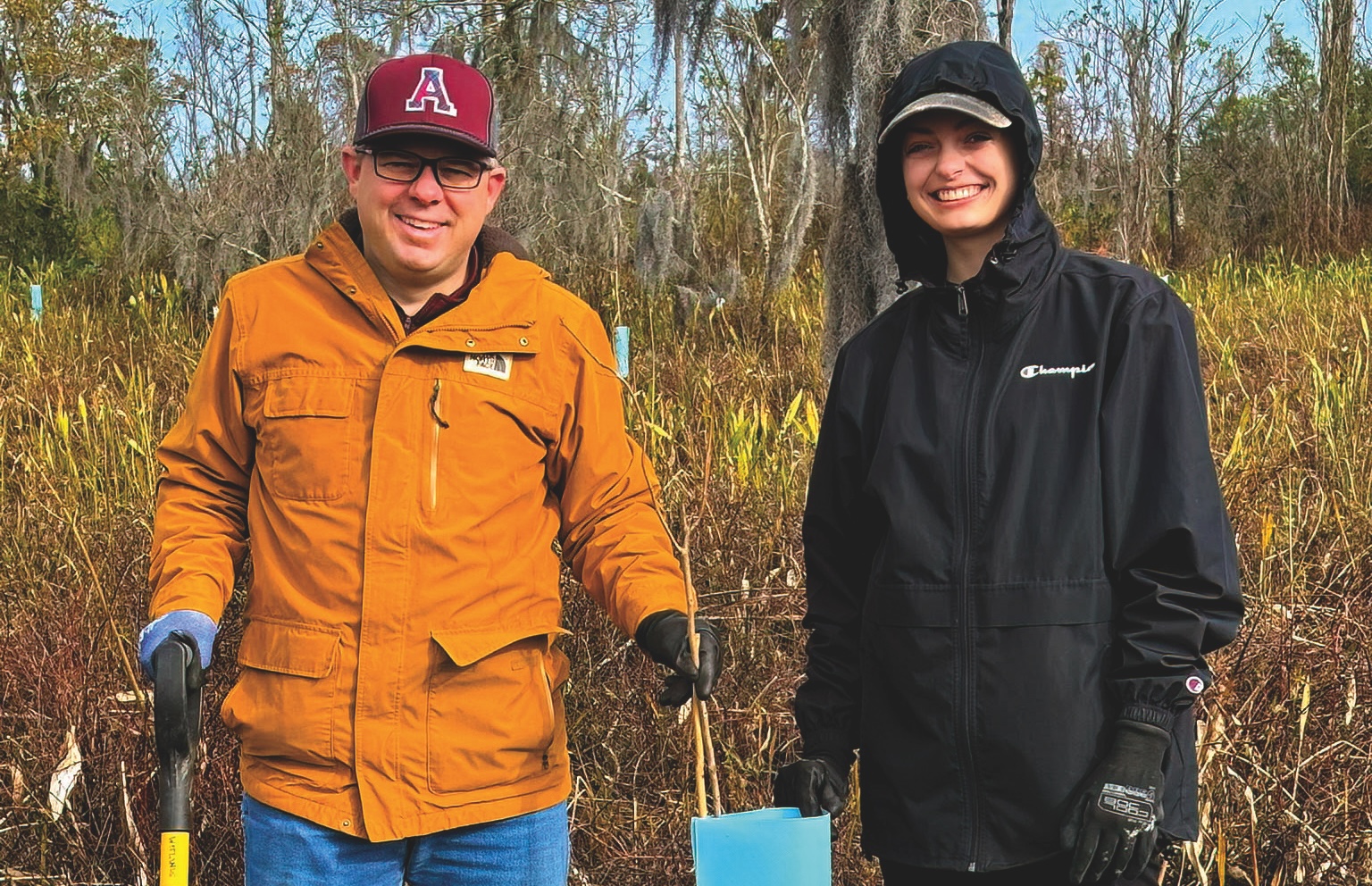 A middle-aged man and a female college student are pictured in an outdoors environment, wearing cold-weather clothes and holding maintenance supplies.