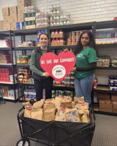Two people stand in a pantry filled with food shelves, holding a large red heart-shaped sign reading “We Love Our Community.” In front of them is a cart filled with paper bags of food.