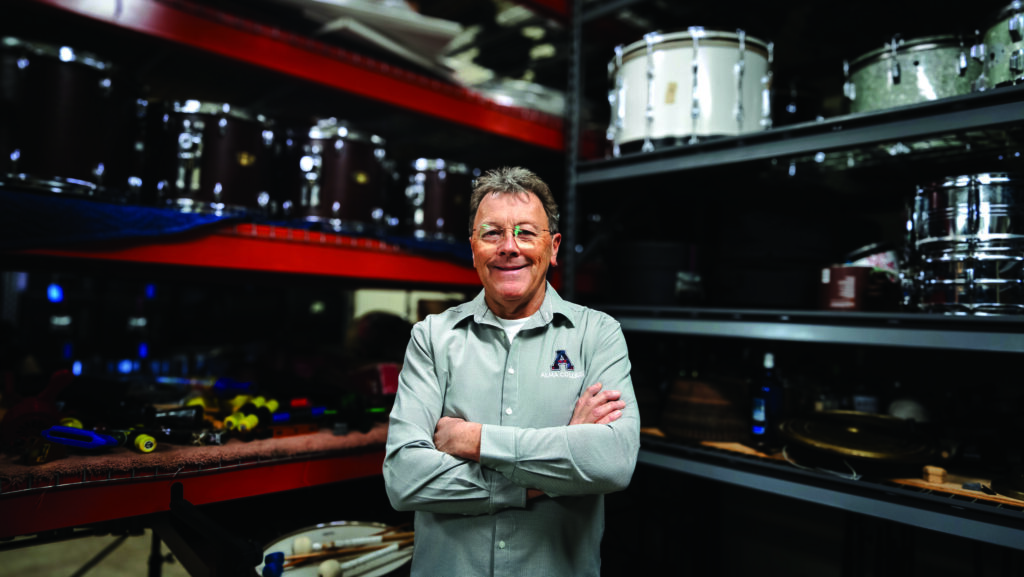 A middle-aged man in a collared shirt stands before a large assortment of marching band equipment being kept in storage.