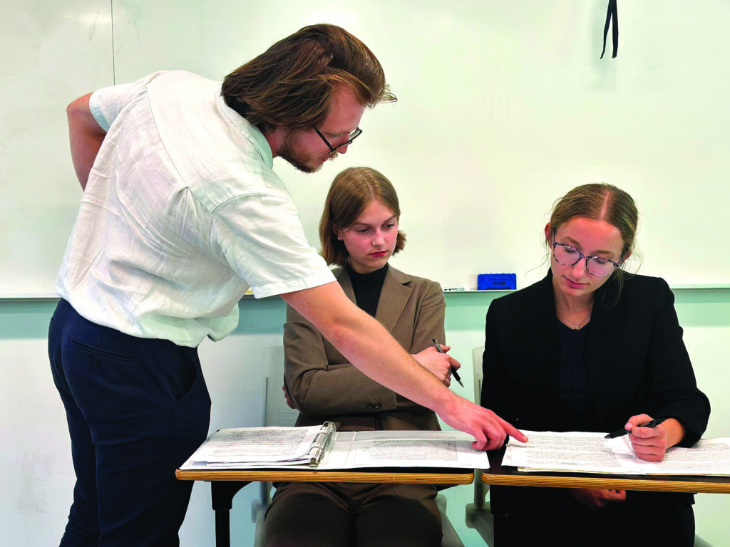 A young male college professor stands near two seated female students and guides them in their lessons.