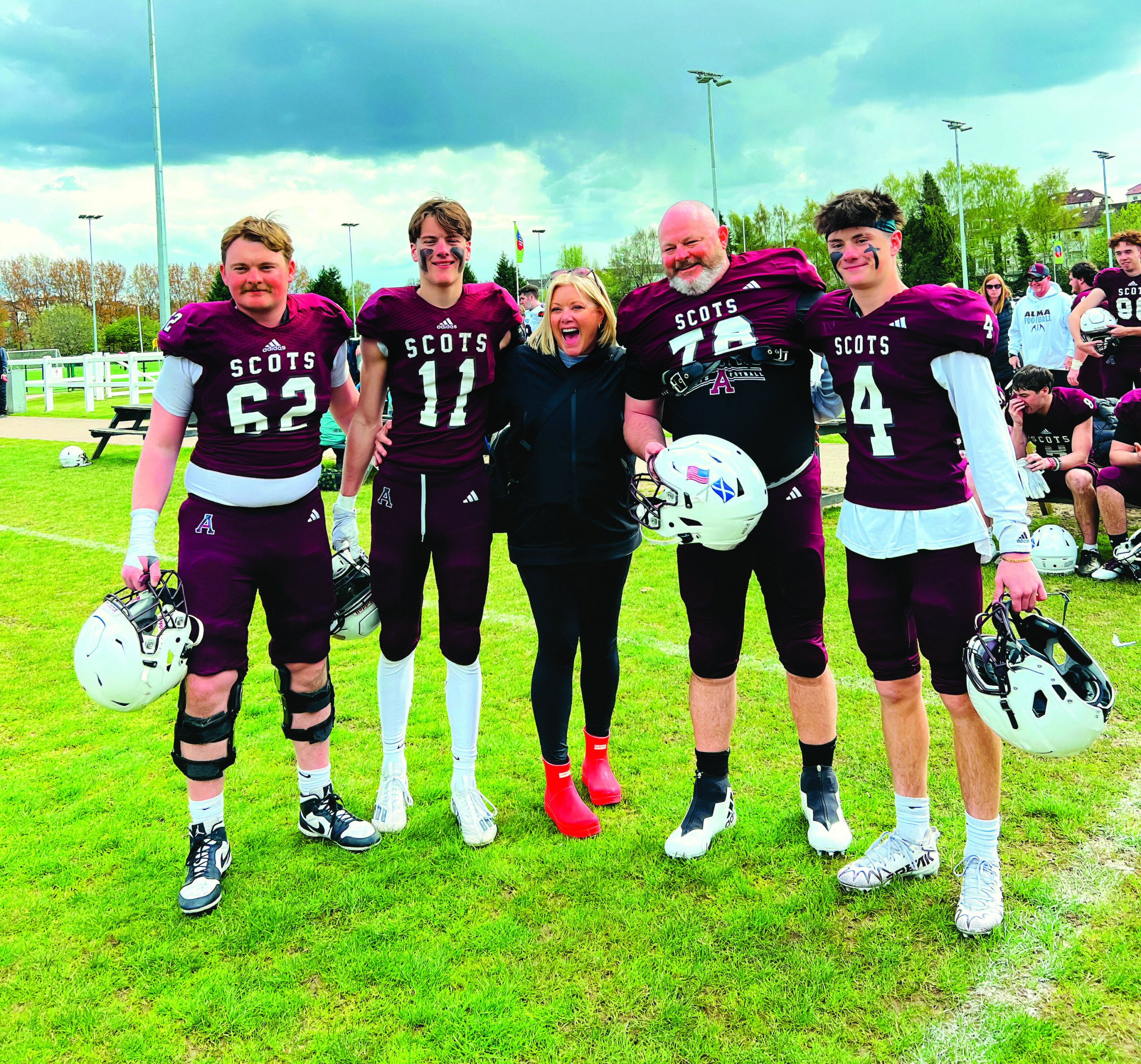 A middle-aged, blonde-haired woman with a big smile stands in the middle of a row of football players: her three sons and husband, who are wearing maroon-colored uniforms and holding their helmets at their sides.