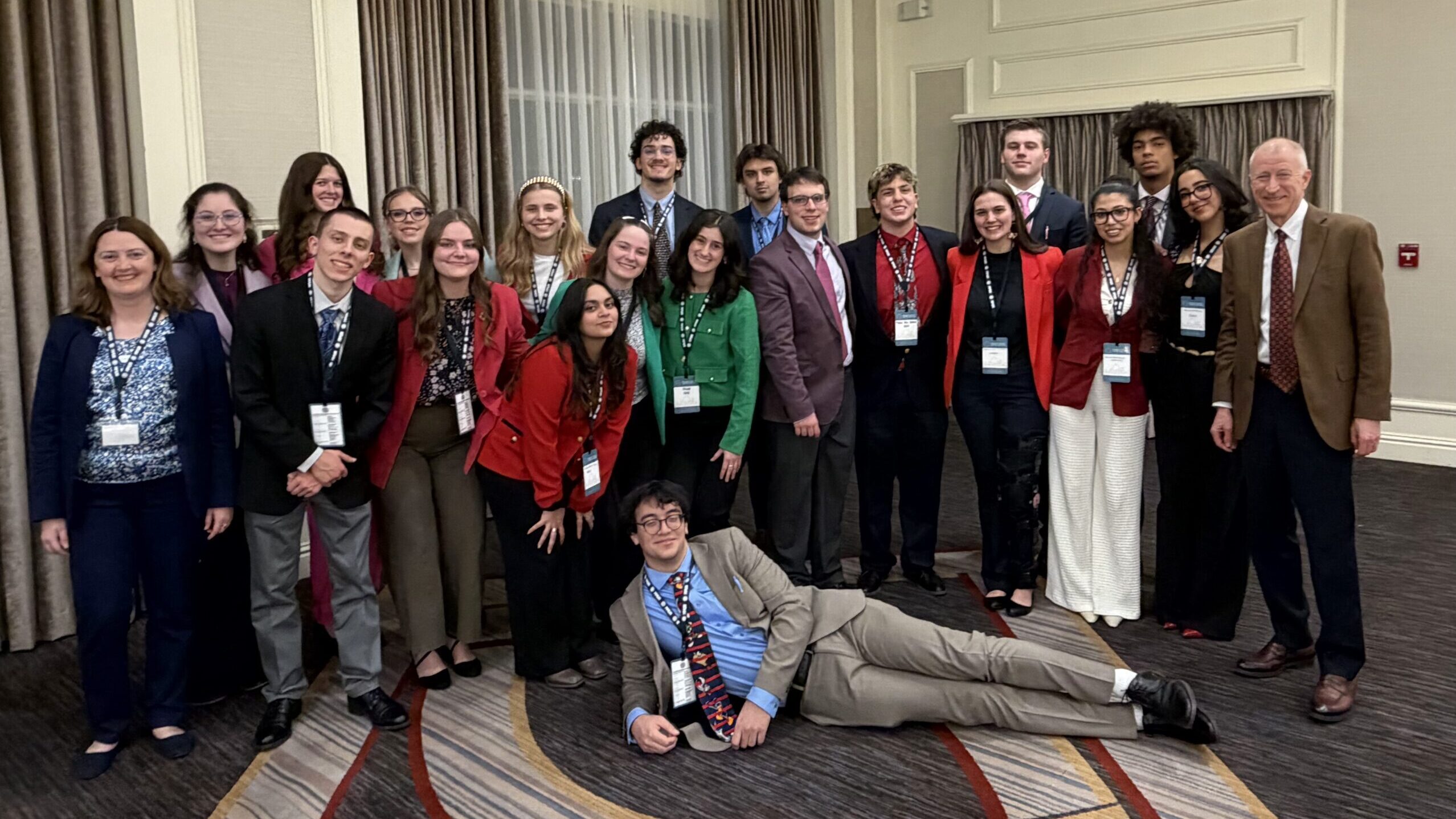 A group of about twenty young adults and two adults pose indoors, dressed in business attire with conference badges. One person is lying playfully in front, while the others stand and smile for the photo.