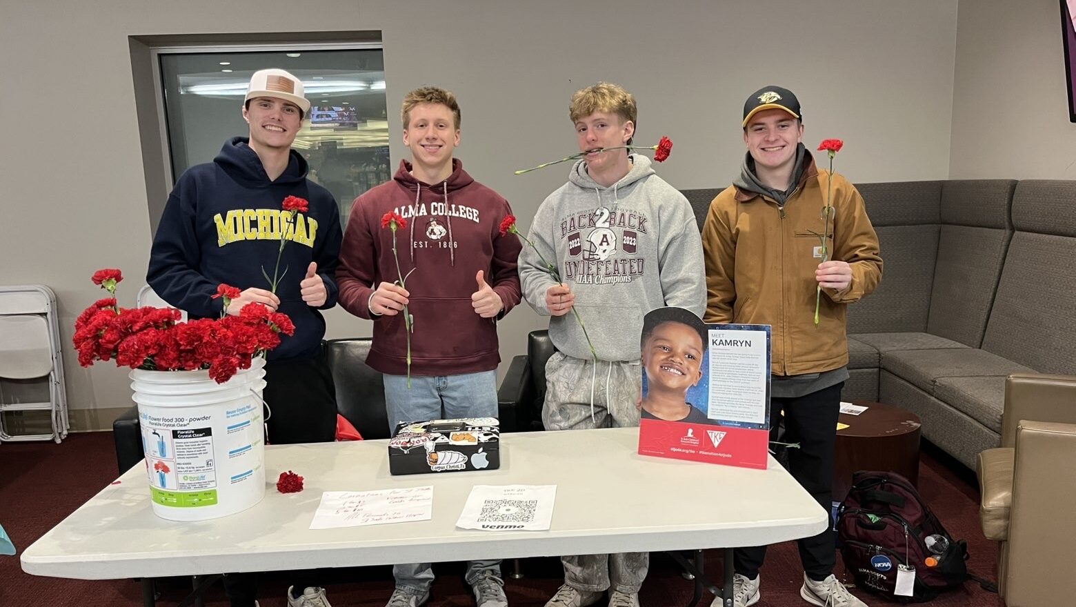 Four young men stand behind a table with red carnations, posing and smiling. The table has a bucket of flowers, a framed photo and info sheet, and a QR code. Some are holding or biting flowers. They appear to be at an indoor event.