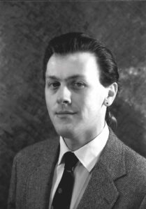 In this photo from the late 1980s, a young man with long hair, dressed in a dark suit, poses for a photo.
