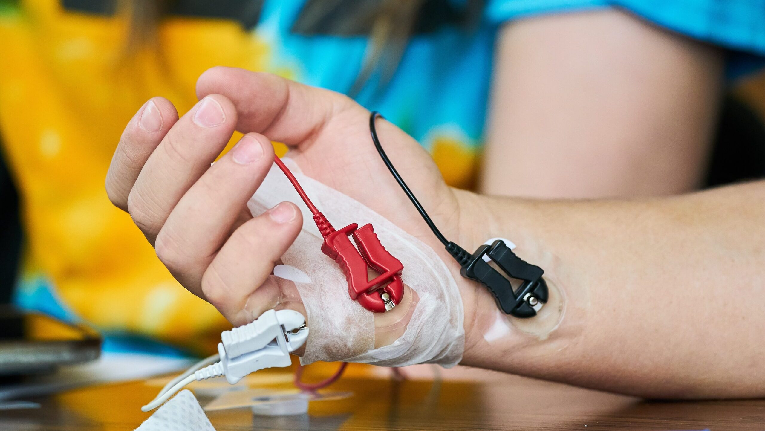 A close-up of a person’s hand and wrist with three medical electrodes, one red, one white, and one black, attached to the skin with adhesive tape. The background is blurred and colorful.
