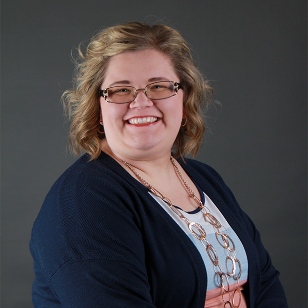 Smiling woman with wavy blonde hair, wearing glasses, a navy blue cardigan, a white and peach top, and a long chain necklace—Kathlyn Allmacher is posed in front of a plain dark background.