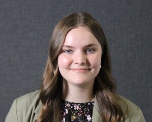 A young woman with long brown hair and blue eyes smiles at the camera. She is wearing a green blazer over a floral top, and stands in front of a plain gray background.
