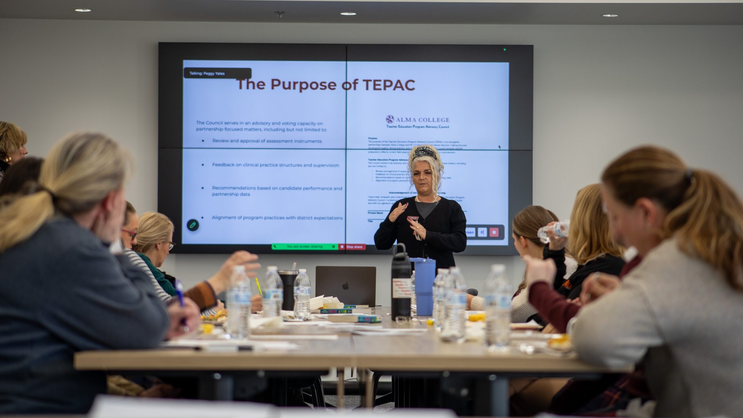 A woman stands and speaks in front of a presentation slide titled The Purpose of TEPAC to a group of people seated at a table with laptops, drinks, and notebooks in a meeting room.