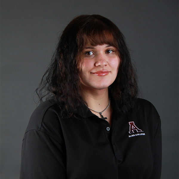 Judith Brooks, with wavy dark hair and bangs, wears a black collared Alma College shirt and smiles slightly against a plain dark background.