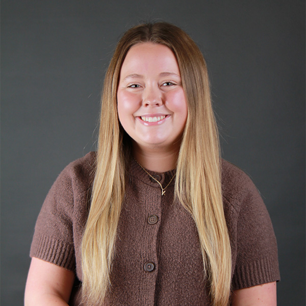 Kathlyn Allmacher, a woman with long, straight blonde hair, wears a brown short-sleeved cardigan and a gold necklace, smiling in front of a plain dark gray background.