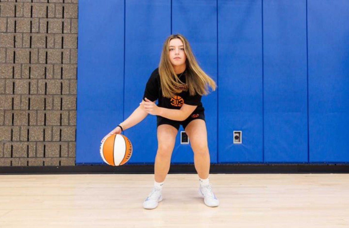 An Alma College student in sportswear dribbles a basketball on an indoor court, bending her knees and focusing ahead. A blue padded wall and a brick wall form the backdrop.