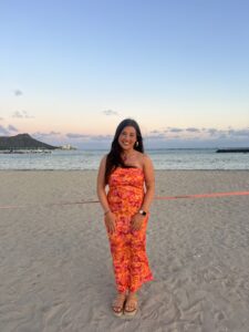 A woman in a colorful orange and pink strapless dress, reminiscent of Alma College students' vibrant spirit, stands smiling on a sandy beach at sunset, with calm ocean waves and a distant mountain in the background.