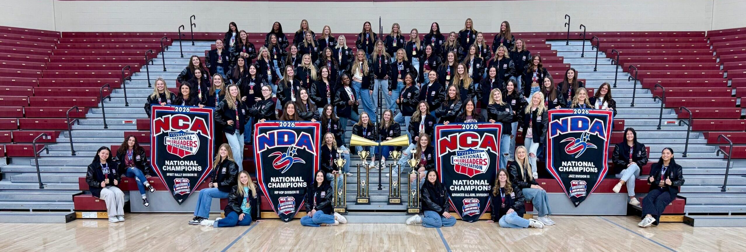 A large group of alma-college-dance-cheer team members pose in bleachers holding trophies and banners. Text above reads 2026 Alma College Cheer & Dance National Champions.
