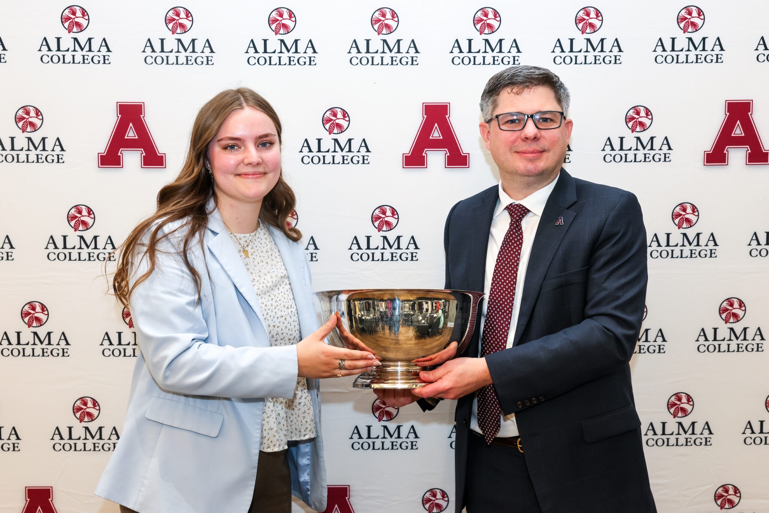 A young woman and a man in a suit smile while holding a large silver trophy together in front of a backdrop with the Alma College logo and red A letters.