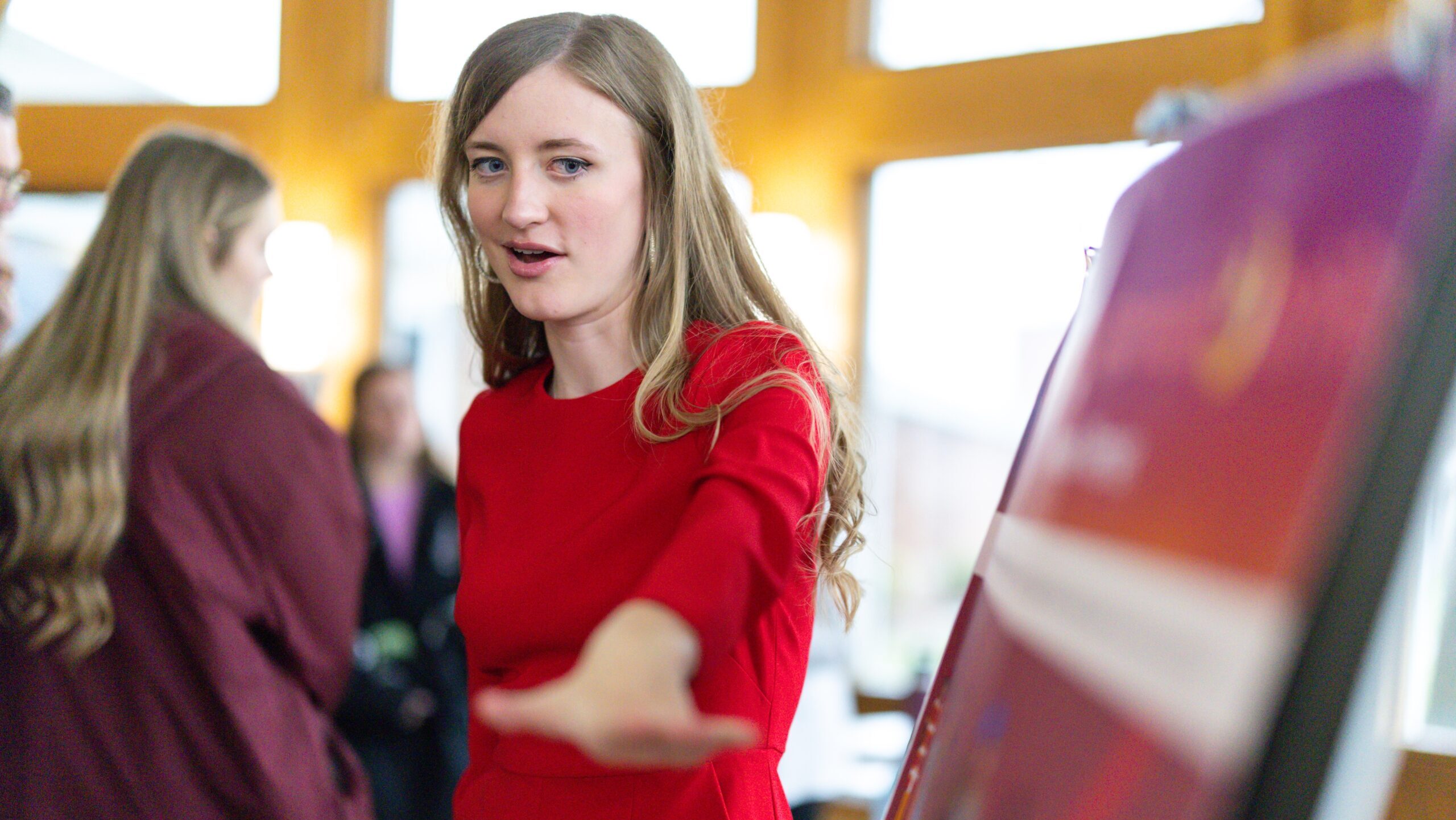 A woman in a red dress gestures toward a display board while speaking, with blurred people and bright windows in the background.