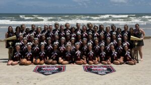 A large cheerleading team poses together on a sandy beach in front of the ocean, wearing matching uniforms with maroon and white bows. Two championship banners are displayed on the sand in front of the group.
