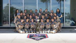 A cheerleading team in matching tan and blue uniforms poses outside a building with their coach, smiling behind a large trophy and an NCA National Champion banner on the ground. Palm trees are visible in the background.