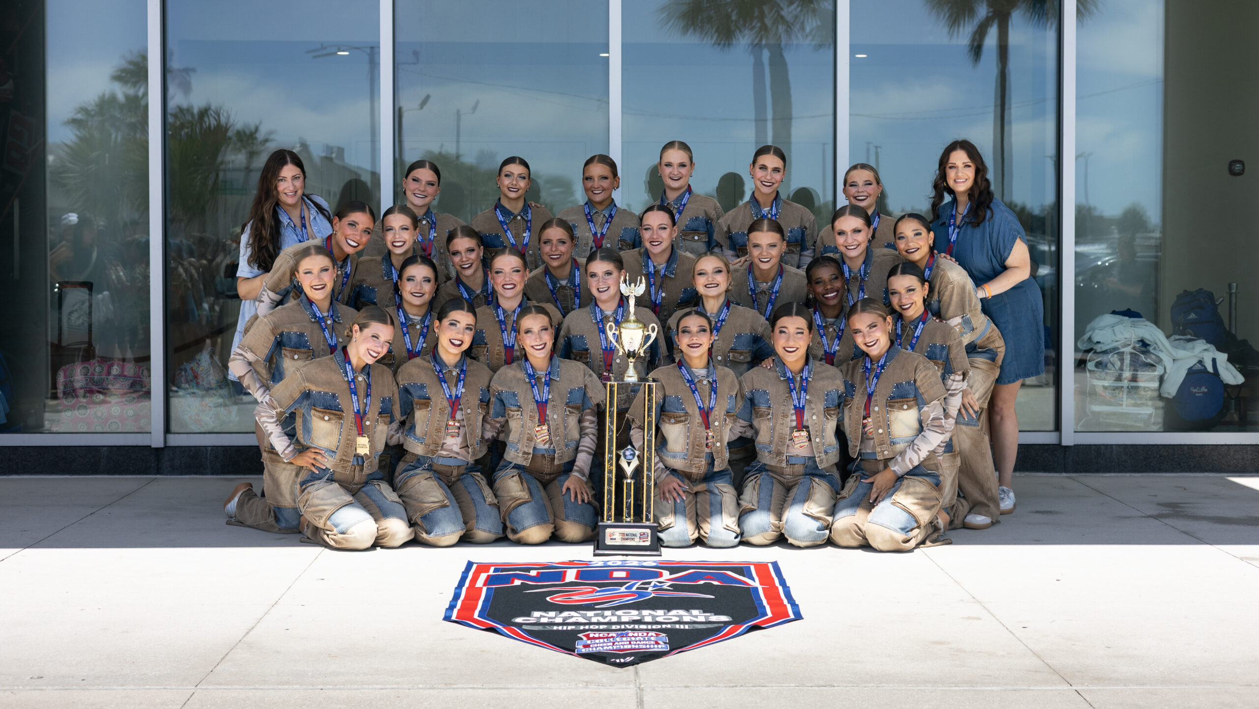 A cheerleading team in matching tan and blue uniforms poses outside a building with their coach, smiling behind a large trophy and an NCA National Champion banner on the ground. Palm trees are visible in the background.