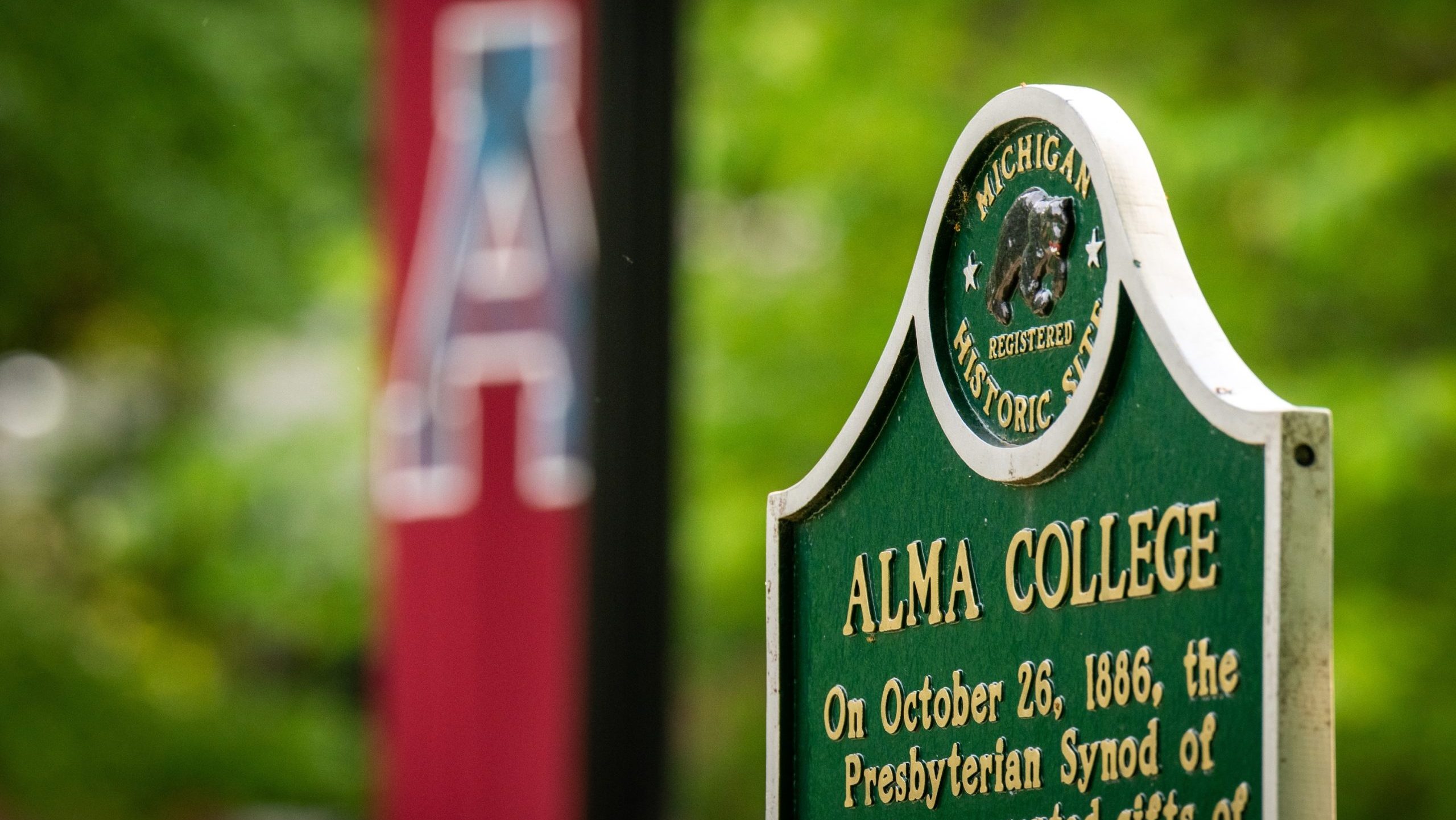 A green Michigan historic site sign for Alma College stands in focus, with a blurred red banner featuring a large white A in the background and lush green trees surrounding them.