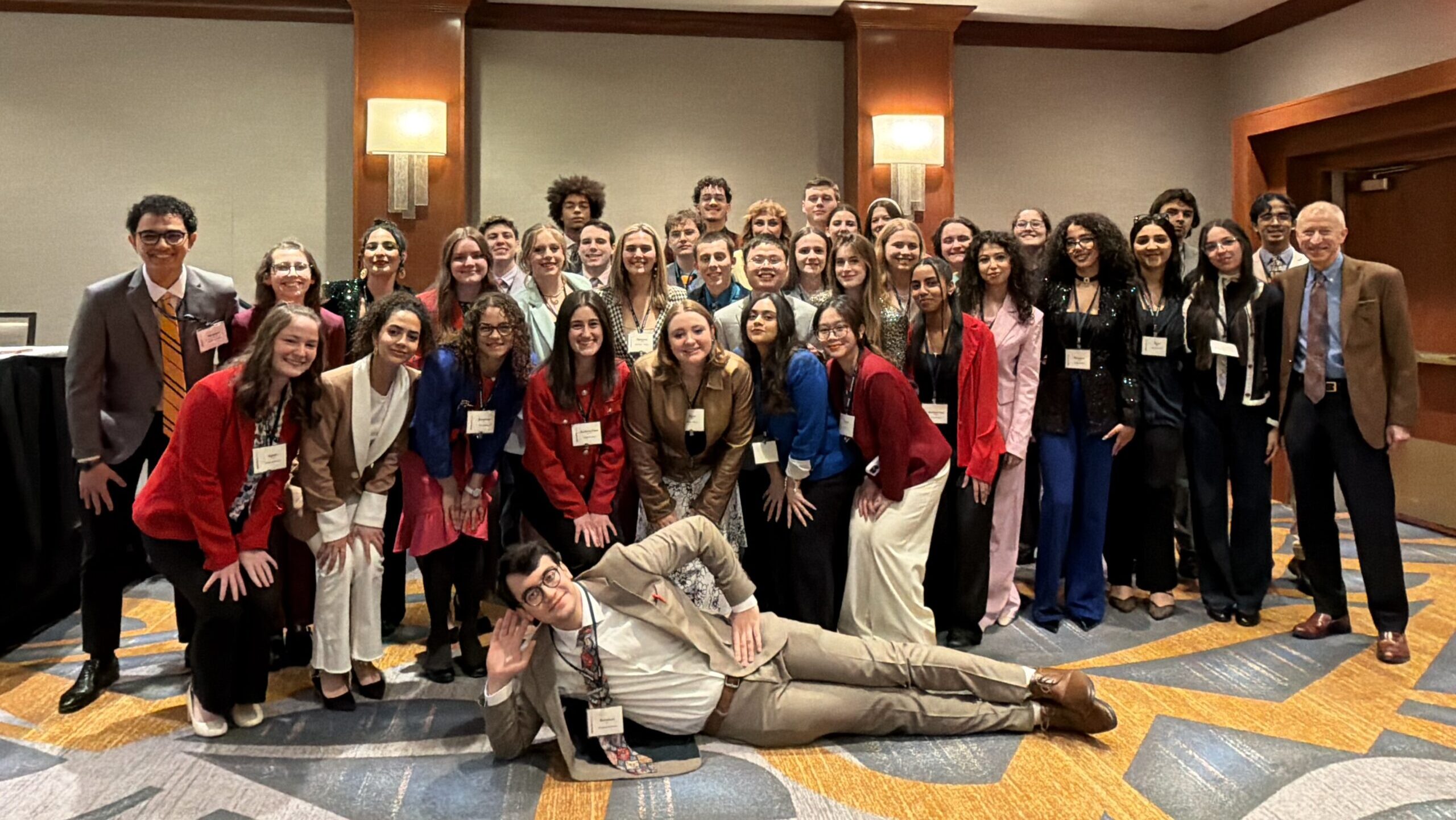 A large group of young adults and a few older adults, dressed in business or formal attire, pose together and smile for a group photo in a carpeted conference room. One person is lying playfully in front.