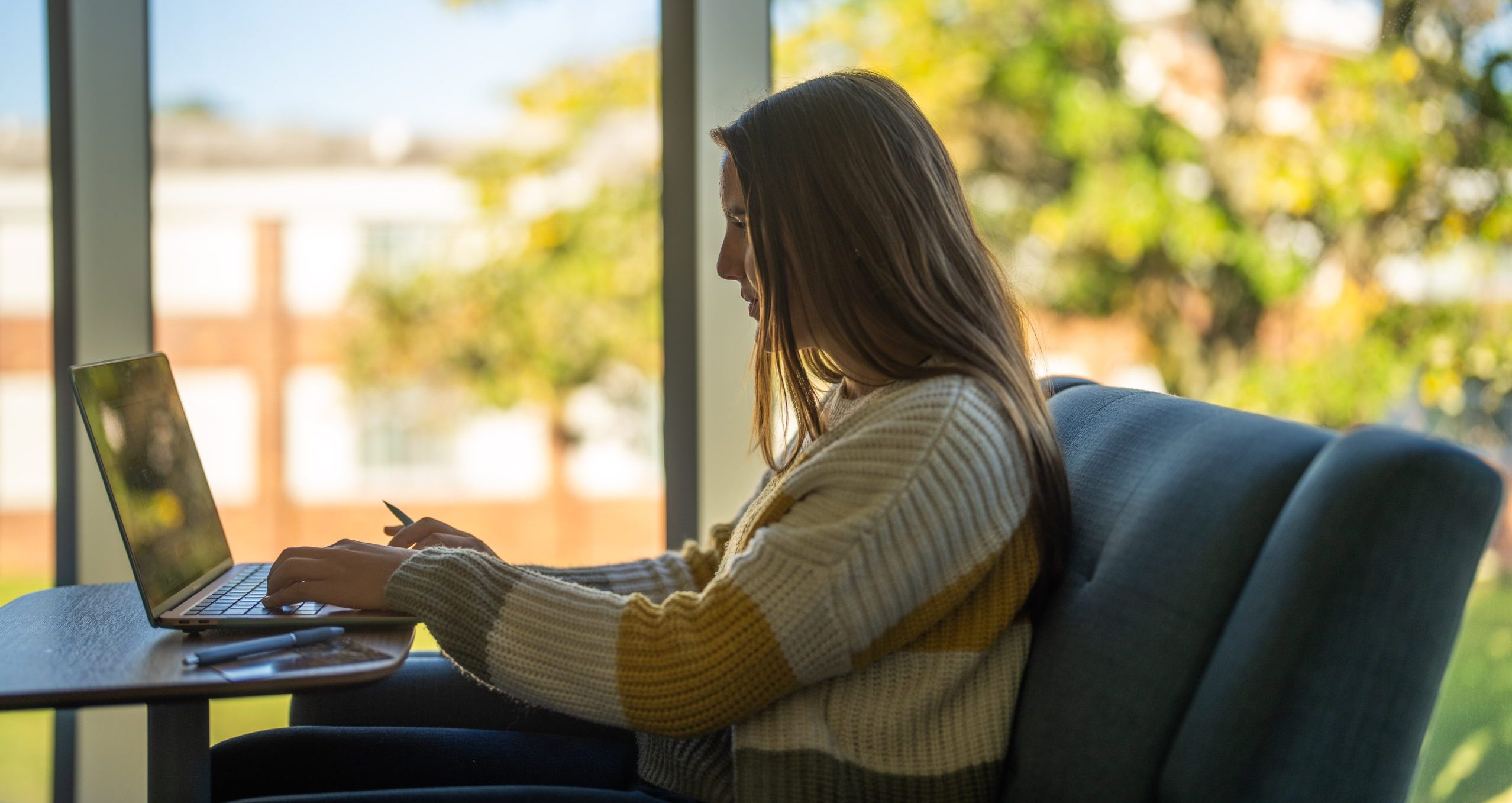 A person sits in a chair by a large window, working on a laptop with Alma open. Sunlight streams in, and trees with green and yellow leaves are visible outside. The person wears a striped sweater and appears focused on the screen.