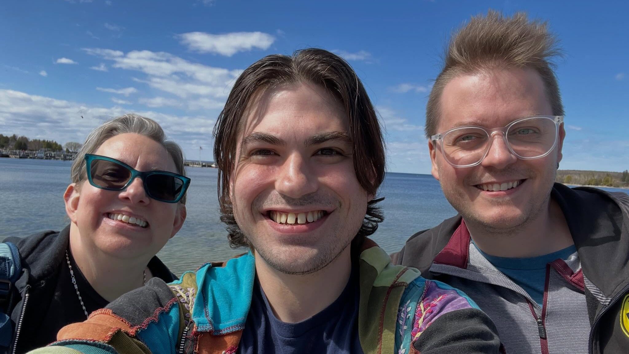 Three MFA graduates smiling and posing for a selfie outdoors near a body of water under a blue sky with some clouds. Trees line the distant shore in Door County. The group appears happy and relaxed, enjoying their writing residency together.