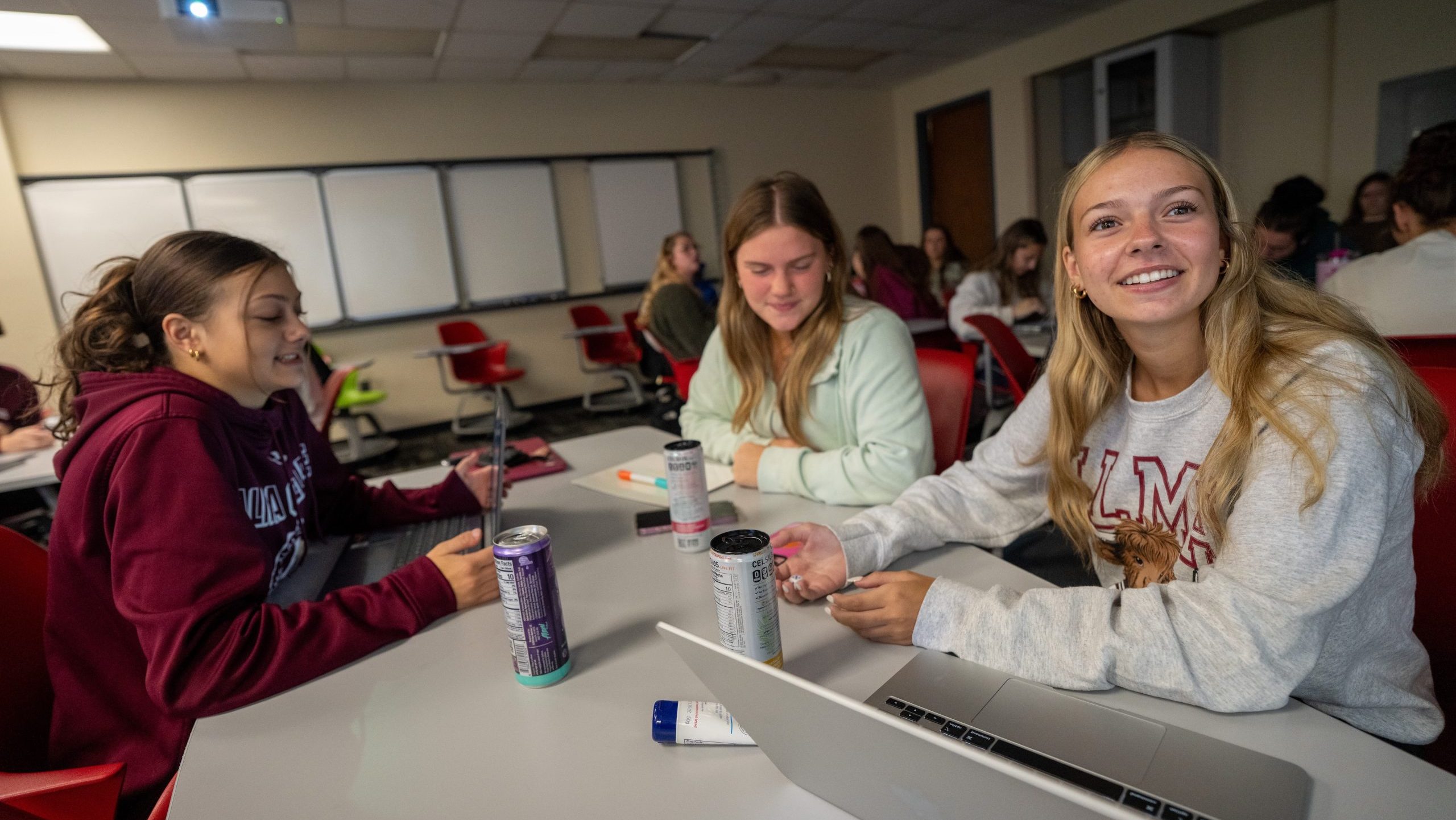 Three students sit at a classroom table at Alma College, smiling and talking. They have laptops, notebooks, and energy drinks in front of them—ready for summer learning. Other students are visible in the background by whiteboards and red chairs.