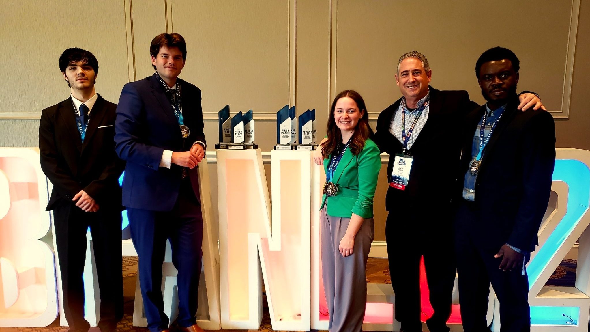 Five BPA students from Alma College stand smiling in formal attire before large, lit-up letters and several trophies, proudly wearing medals—celebrating their impressive national finishes at an awards or recognition event.