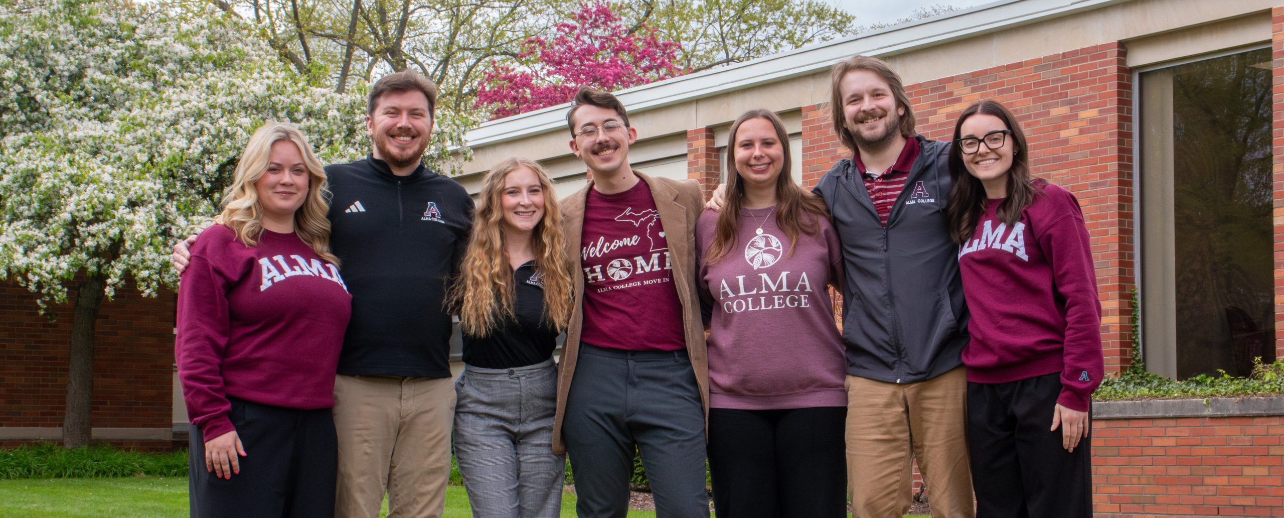 Seven people stand side by side smiling outdoors in front of a brick building, many wearing Alma College sweaters or shirts. Trees with white and pink blossoms bloom in the background—connect with your admissions team and picture yourself here!