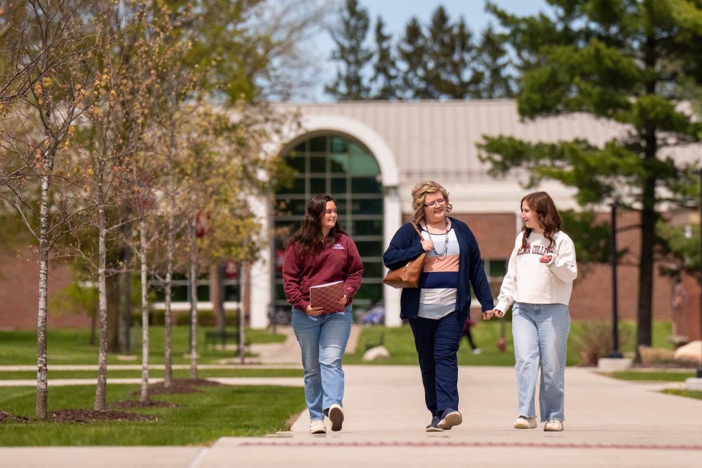 Three women walk together on a campus pathway, talking and smiling. As they connect-and-visit, trees and a building with large windows are in the background. Two women hold books or notebooks, enjoying the pleasant weather.