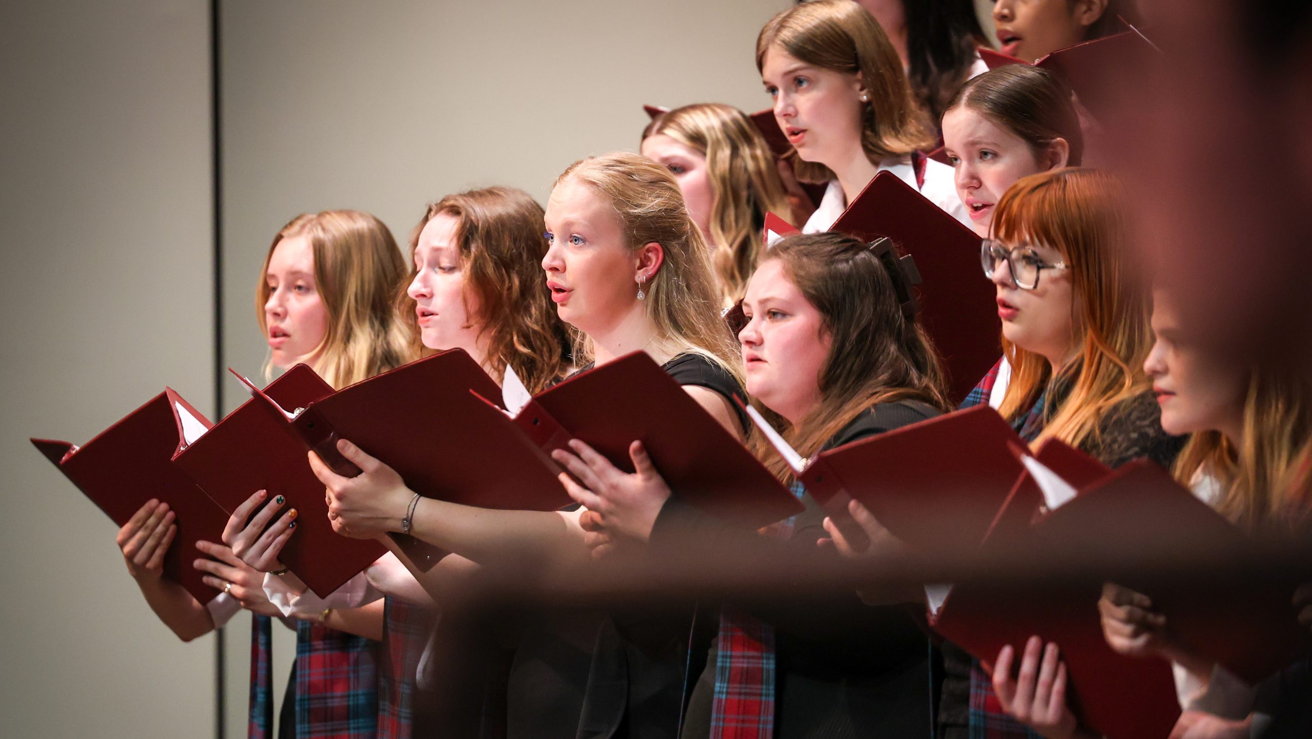A group of young women in matching plaid uniforms perform a choir piece at Alma College, holding open red folders with sheet music, as they debut a new work by composer Justin Rito.